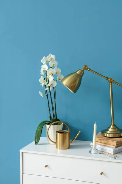 Orchid flower, watering can, lamp and books on chest of drawers near color wall