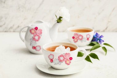 Stylish tea pot and cups of hot beverage with flowers on light background
