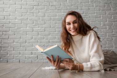 Gri tuğla duvarın yanında, ahşap zeminde kitap okuyan güzel genç bir kadın.