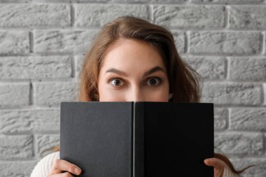 Young woman holding opened book near grey brick wall