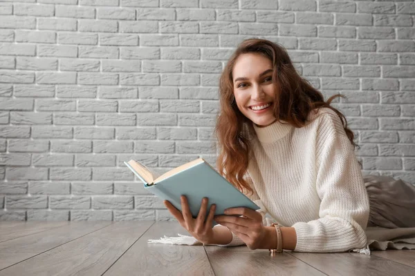 Gri tuğla duvarın yanında, ahşap zeminde kitap okuyan güzel genç bir kadın.