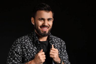 Gülümseyen sakallı adam koyu arkaplanda gömlek ayarlıyor.