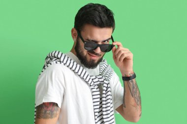 Güneş gözlüğü takan ve yeşil arka planda göz kırpan modaya uygun sakallı bir adam.