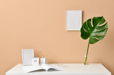 Boş fotoğraf çerçeveleri, not defteri ve bej duvar kenarındaki tropikal yapraklı vazo ile şık bir iş yeri.