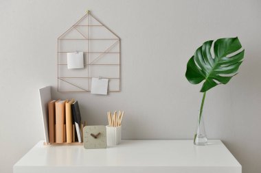 Stylish workplace with books, clock and vase with tropical leaf near light wall