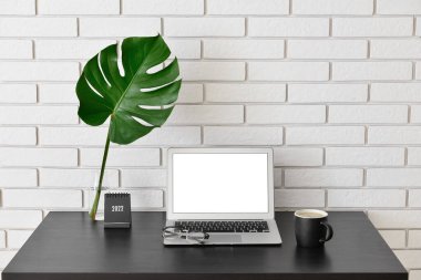 Laptoplu modern işyeri, bir fincan kahve ve beyaz tuğla duvarın yanında tropikal yapraklı vazo