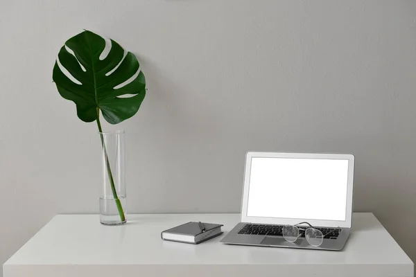 Modern workplace with laptop, notebook and vase with tropical leaf near light wall