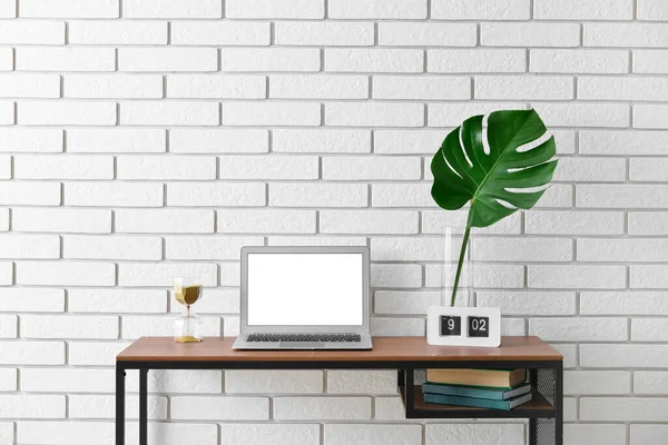 Beyaz tuğla duvarın yanındaki vazoda dizüstü bilgisayarı, kum saati, saati ve palmiye yaprağı olan işyeri