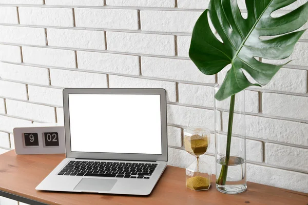 Beyaz tuğla duvarın yanındaki vazoda dizüstü bilgisayarı, kum saati, saati ve palmiye yaprağı olan işyeri