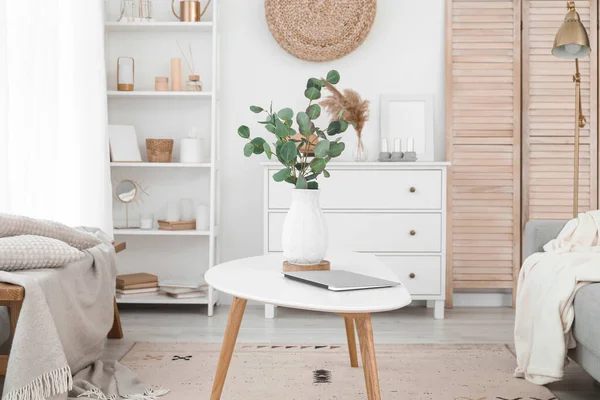 Vase with eucalyptus branches and laptop on table in light living room
