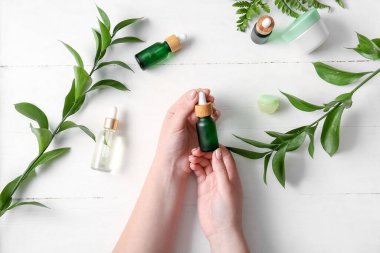 Doğal serumdan şişeler, kozmetik ürünleri ve hafif ahşap arka planda bitki dalları olan kadın eller.