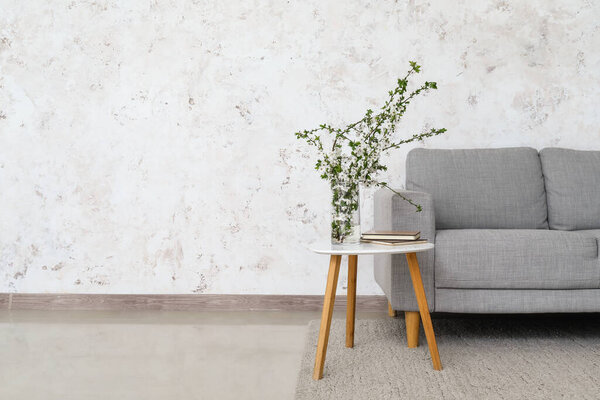 Vase with blooming branches and books on table near sofa in light room
