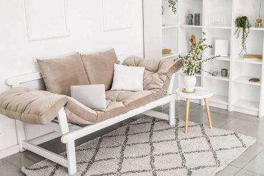 Dizüstü bilgisayarlı kanepe, vazoda çiçek açan dallar masanın üstünde ve ışık duvarının yanında raf ünitesi.