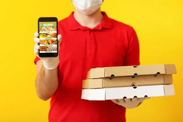 Male courier of food delivery company in medical mask, with mobile phone and tasty pizza on yellow background