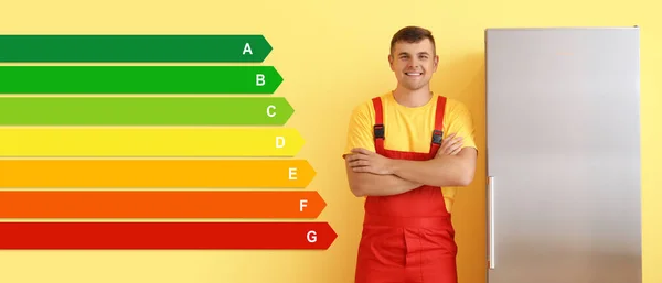 Worker of repair service near modern refrigerator and energy efficiency rating on yellow background. Concept of smart home