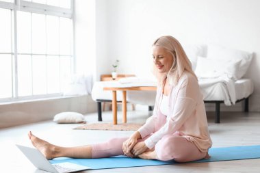 Bilgisayarlı olgun bir kadının evde yoga yaptığı bir sabah.