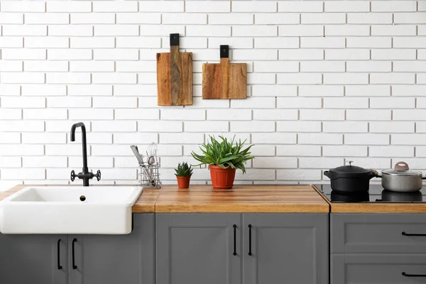 Kitchen with stylish furniture, sink, houseplants and utensils near white brick wall