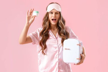 Pijamalı güzel bir kadın ve pembe arka planda kozmetik ürünleri için küçük bir buzdolabı var.