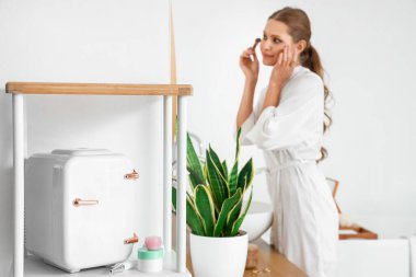 Kozmetik ürünleri için küçük bir buzdolabı ve evde makyaj yapan güzel bir kadın.