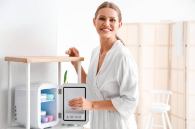 Evdeki kozmetik ürünleri için küçük buzdolabından krema alan güzel genç bir kadın.