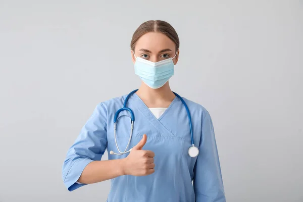 灰色の背景に親指アップを示す医療用マスクの女性看護師 — ストック写真