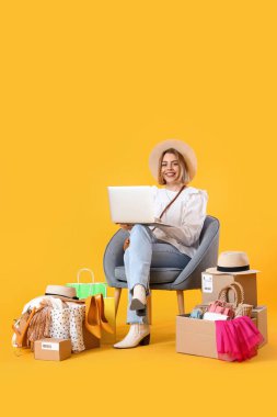Sarı arka planda dizüstü bilgisayarı ve alışveriş çantaları olan genç bir kadın.