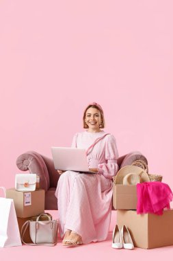 Dizüstü bilgisayarı ve yeni kıyafetleri olan genç bir kadın pembe arka planda koltukta oturuyor. İnternetten alışveriş