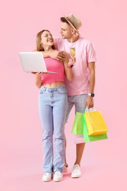 Laptopu, kredi kartı ve pembe arka planda alışveriş torbaları olan genç bir çift. İnternetten alışveriş