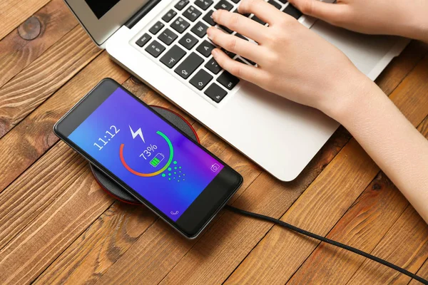 Mobile phone charging on wireless pad and woman using laptop on wooden background