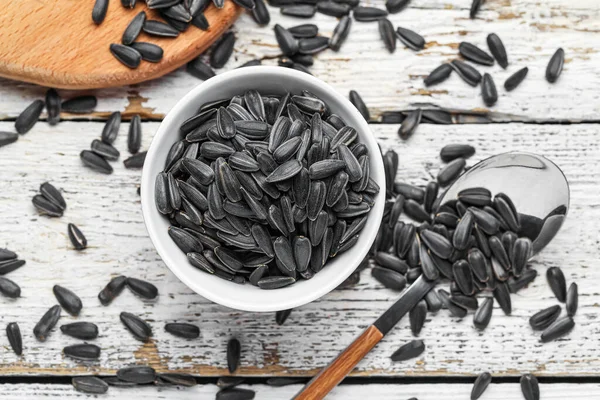 Bowl and spoon with black sunflower seeds on white wooden background