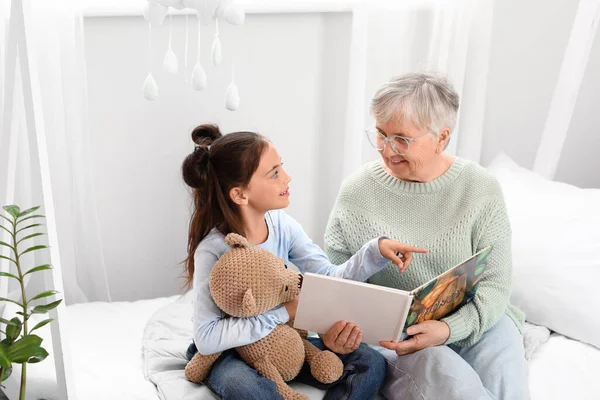 Little girl with toy and her grandma reading book in bedroom