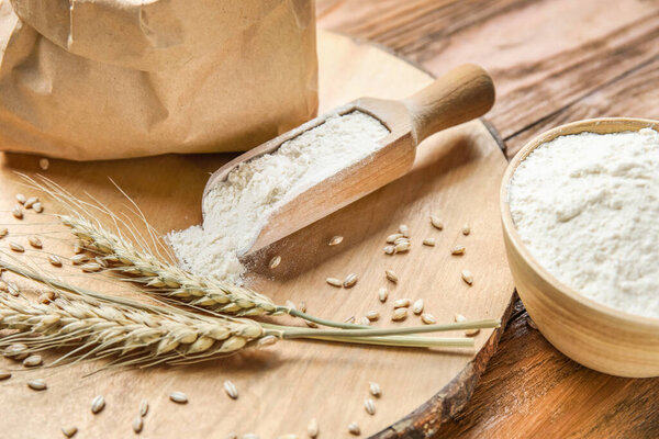 Пшеничные уши, бумажный пакет, совок и миска с мукой на деревянном фоне, крупным планом