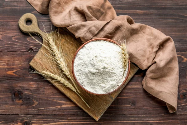 Board, bowl with flour, wheat ears and napkin on dark wooden background