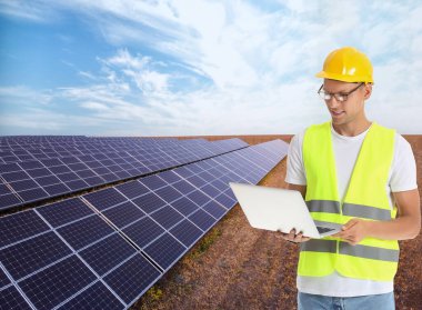 Modern güneş enerjisi santralinin yakınında, kasketli ve dizüstü bilgisayarı olan genç bir erkek mühendis.
