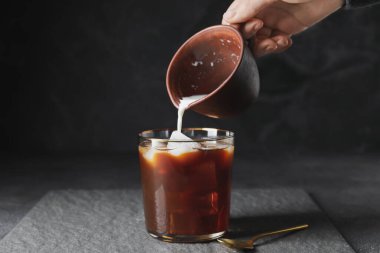 Kadın koyu arkaplanda soğuk kahve bardağına süt döküyor.