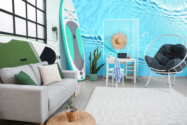 Interior of living room with comfortable furniture, workplace and sup surfing board near wall with print of clear water