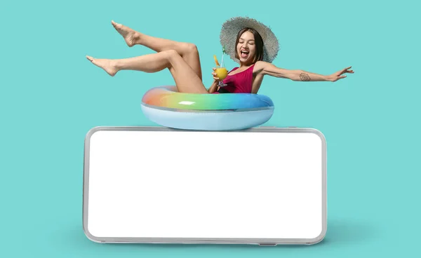 Happy young woman with cocktail, inflatable ring and mobile phone with blank screen on color background