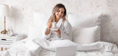 Laptoplu güzel bir kadın yatak odasında kahve içiyor.