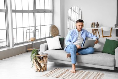 Kulaklık ve dizüstü bilgisayarlı genç çıplak ayaklı adam evdeki kanepede oturuyor.