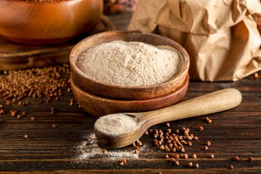 Koyu ahşap arka planda bir kase un, kaşık ve karabuğday taneleri.