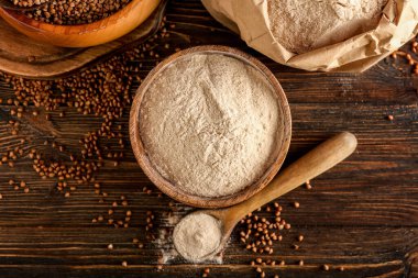 Koyu ahşap arka planda bir kase un, kaşık ve karabuğday taneleri.