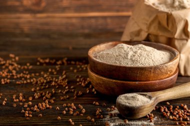 Koyu ahşap arka planda bir kase un, kaşık ve karabuğday taneleri.