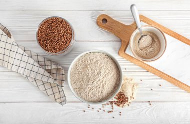 Beyaz ahşap arka planda bir kase un, karabuğday taneleri, peçete ve tahta.