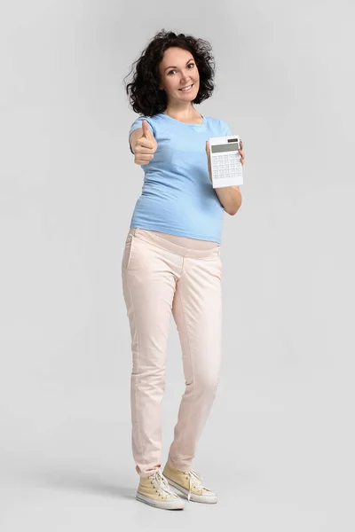 Young pregnant woman with calculator showing thumb-up on light background