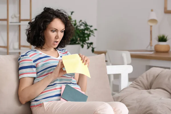 Shocked pregnant woman opening envelope on couch at home