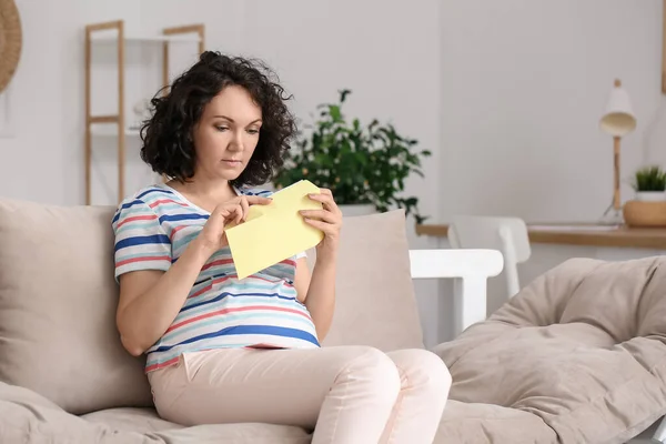 Young pregnant woman opening envelope on couch at home