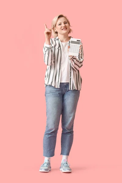 Beautiful blonde woman with calculator pointing at something on pink background