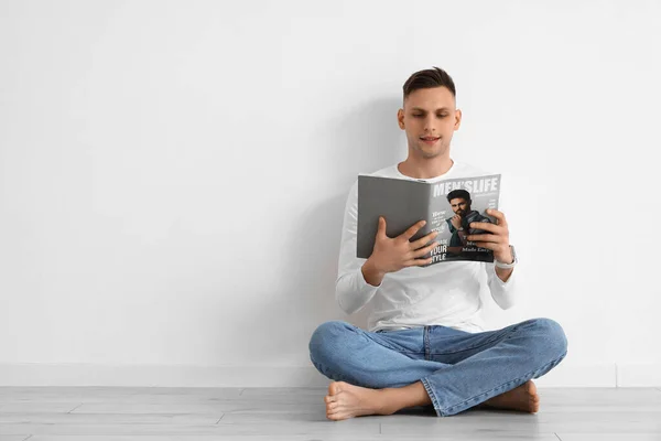 Young barefoot man reading magazine near light wall