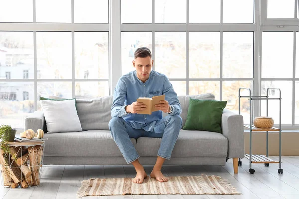 Young barefoot man reading book on sofa at home