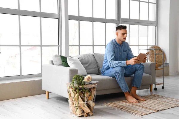 Young barefoot man reading book on sofa at home